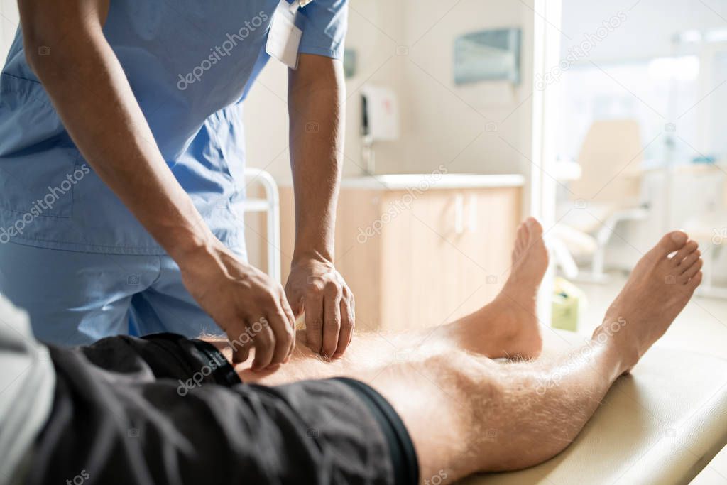 Sick patient lying on couch while clinician in blue uniform bending over him and massaging his leg