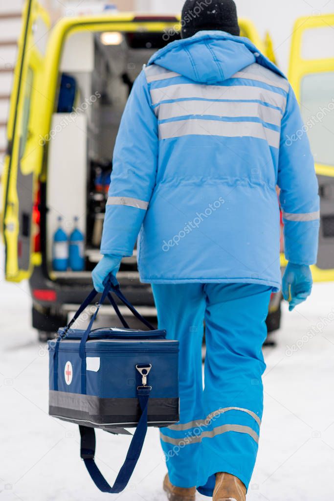 Rear view of paramedic in blue workwear and medical gloves carrying first aid kit while going towards ambulance car