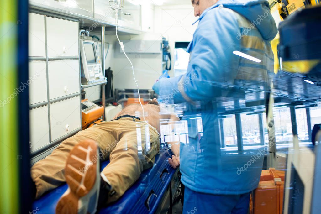 Young paramedic in uniform standing by stretcher with sick shirtless man while giving him first aid inside ambulance car