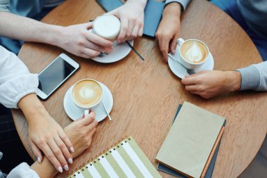 Overview of hands of college students with gadgets and drinks gathered by table in cafe for cup of cappuccino and chat