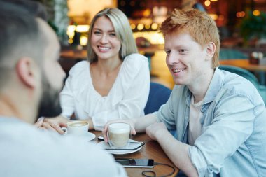 Happy casual guy with cup of cappuccino and his girlfriend looking at their friend during chat by table in cafe