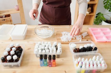 Kahverengi önlüklü genç bir kadın, tahta tahtadan bir parça alıp sabun yaparken kaseye koyuyor.