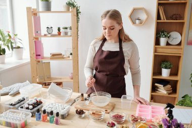 İş kıyafetlerindeki güzel kadın silikon kalıplara dökerken sıvı sabun kütlesini kaseye karıştırıyor.