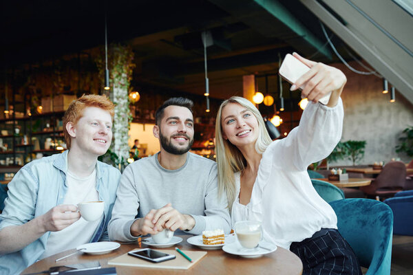 Group of cheerful young friends making selfie in cafe while sitting by table and enjoying rest by cup of cappuccino