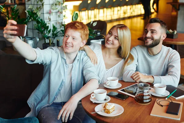 Three happy young affectionate friends looking at smartphone camera while making selfie in cozy cafe by cup of tea