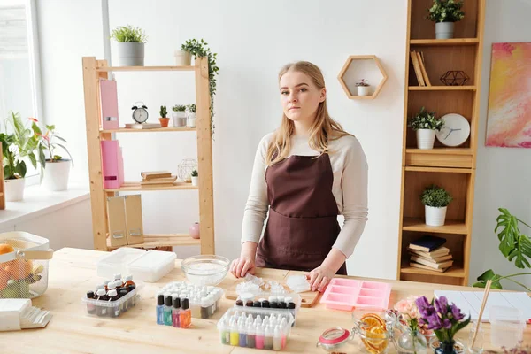 若いですブロンド職人女性で茶色いエプロン立ってテーブルの上にスタジオで作る手作り石鹸 — ストック写真