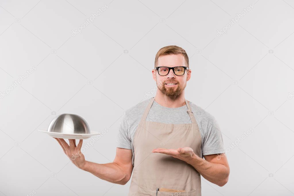 Happy young bearded waiter in apron and eyeglasses showing cloche with cooked food while standing in front of camera in isolation