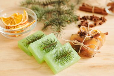 Kozalaklı el yapımı sabunlar, aromatik baharatlar ve ahşap masa üzerinde portakal dilimleri olan cam eşyalar.