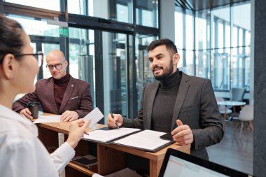 Bearded young businessman with pen looking at female receptionist while filling in form by counter in hotel lounge