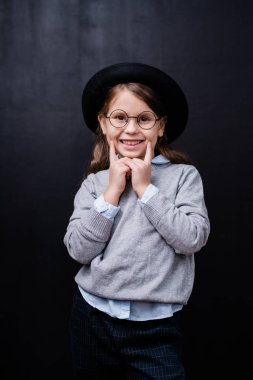 Güzel, küçük bir kız, gözlüklü, günlük kıyafetli, dişlek bir gülümsemeyle kameranın önünde dikilirken sana bakıyor.