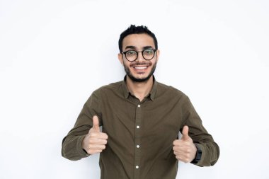 Genç, dişlek gülümseyen, baş parmağını kaldıran, yalnız başına kameranın önünde duran neşeli bir adam.