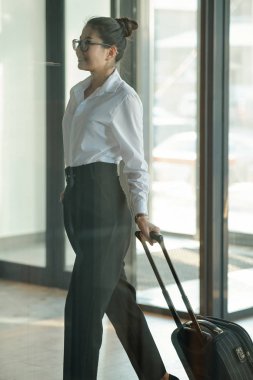 Pretty young Asian businesswoman pulling suitcase with baggage while moving towards elevator after entering hotel