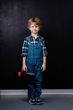 Kot pantolon ve gözlüklü küçük çocuk siyah arka planda tek başına dikilirken alet çantası tutuyor.