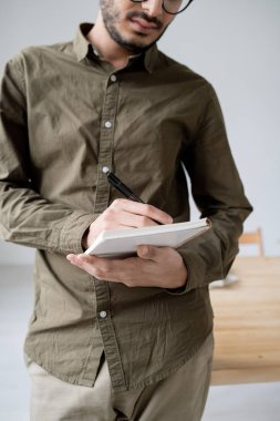 Genç, ciddi bir erkek çalışan, ofiste çalışırken not defterine not alıyor.