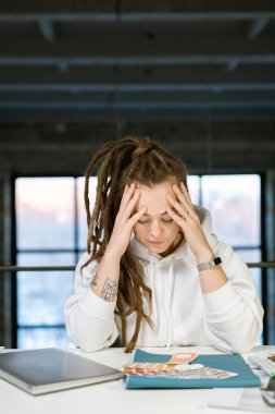 Pensive female designer touching her head while concentrating on work over new fashion collection in studio clipart