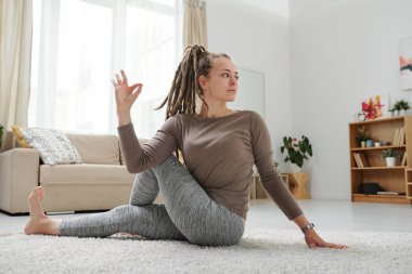 Rastalı genç bir kadın yoga pozisyonunda yerde oturuyor. Kol ve bacak kaslarını esnetiyor.