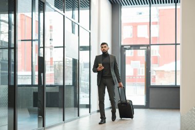 Young elegant businessman with smartphone pulling suitcase with luggage while moving along airport