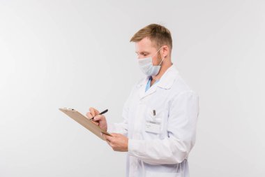 Whitecoat ve koruyucu maskeli genç doktor tecritteki hastaya reçete yazıyor.