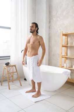 Köpükle sıcak bir banyo yaptıktan sonra kalçasında yumuşak beyaz havluyla küçük bir halının üzerinde duran genç üstsüz adam.