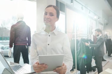 Beyaz gömlekli güzel bir tezgahtar, yeni kıyafetler seçen alıcının arka planında iş yeri teması kullanıyor.