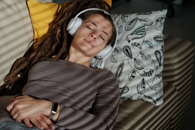 Kulaklıklı, karanlık odada yastığa uzanmış, rahat bir müzik eşliğinde uyuyan genç ve dingin bir kadın.