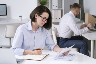 Bayan ofis müdürü, arka planda meslektaşıyla masa başında otururken evrakları karıştırıp not alıyor.