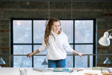 Beyaz kapüşonlu ve kot pantolonlu genç yaratıcı tasarımcı iş yerinde kameranın önünde duruyor.