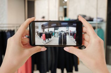 Akıllı telefon tutan ve içinde elbise rafları olan çağdaş bir butiğin fotoğrafını çeken genç bir kadının elleri.