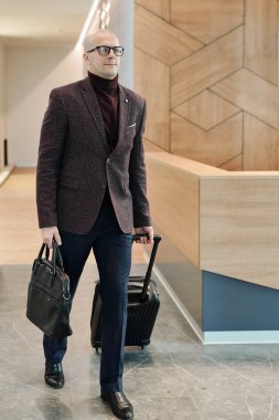 Bald mature businessman with handbag pulling suitcase while moving along reception counter in hotel lounge