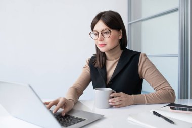 İçki içen genç iş kadını bilgisayarın önünde internetteki finansal verileri analiz ediyor.