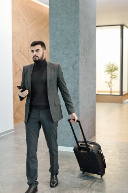 Young businessman with mobile phone pulling suitcase with baggage while moving along hotel lounge
