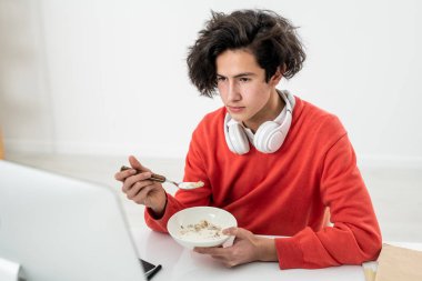 Genç erkek müsli süt içerken bilgisayar monitörünün önünde oturup video izlerken