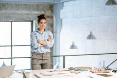 Genç gülümseyen moda tasarımcısı stüdyoda kameranın önünde beklerken kollarını göğsünde kavuşturuyor.