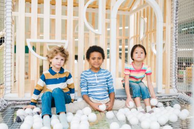 Çeşitli etnik kökenlere sahip üç tatlı çocuk oyun parkında beyaz balonlarla oynuyor.