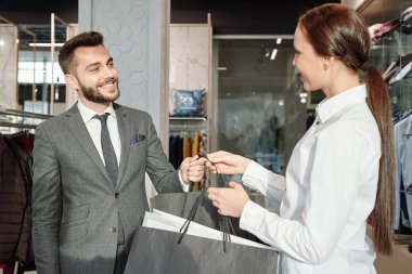 Dost canlısı genç tezgahtar, yeni kıyafetlerle birlikte butikte şık takım elbiseli mutlu iş adamına kağıt torbaları uzatıyor.