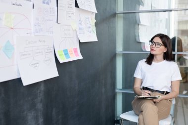 Güzel genç öğrenci ya da iş kadını sandalyede oturup tahtadaki kağıtlara bakarken notlar alıyor.