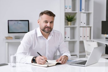 Kendine güvenen yetişkin iş adamı, çalışma planını not defterine yazarken kameranın önünde masasının başında oturuyor.