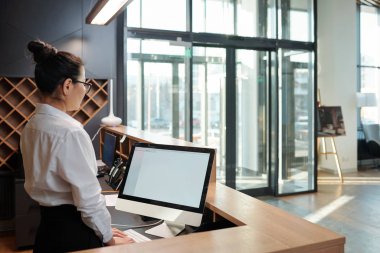 Young elegant female receptionist standing in front of computer screen by counter and waiting for guests in hotel