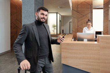 Elegant business traveler with suitcase and smartphone standing in hotel lounge on background of working receptionist