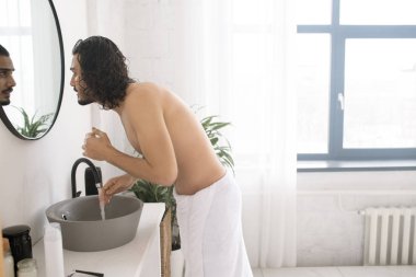 Banyo lavabosunda yüzünü yıkarken kalçasında beyaz havlu olan gömleksiz genç adam aynaya bakıyor.