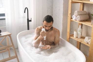 Banyonun köşesinde köpükle banyo yaparken duş jeli kullanan ıslak genç adam.