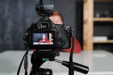 Video camera with young male vlogger or photographer with photocamera on screen during shooting in studio clipart