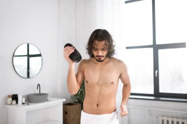 Kalçasında beyaz havlusu olan enerjik bir adam ve banyoda dans eden taşınabilir kablosuz bluetooth kulaklığı.