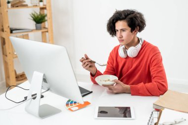 Ciddi genç serbest çalışan bilgisayar monitörünün önünde oturuyor, video izliyor ve öğle arasında sütle müsli yiyor.