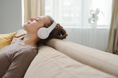 Evde rahat bir kanepede otururken dinlenmek için müzik dinleyen, kulaklıklı, dingin, genç bir kadın.