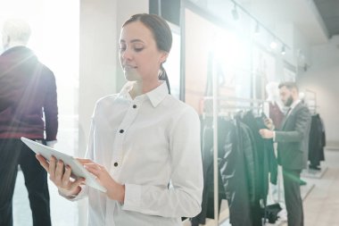 Beyaz gömlekli genç, zarif mağaza asistanı yeni koleksiyon arayan müşterinin arka planında dokunmatik kaydırma yapıyor.