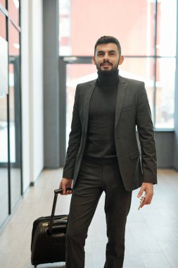 Young businessman with suitcase and smartphone standing in front of camera while waiting for departure announcement in airport
