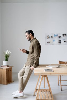 Ofis penceresinin önünde masanın önünde dururken akıllı telefondan kayan gündelik giyimli genç bir iş adamı.