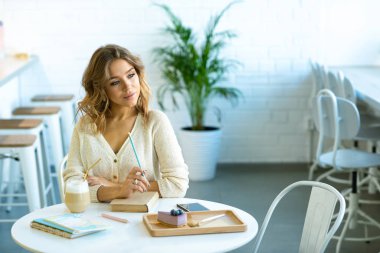 Güzel sarışın öğrenci, kafeteryada masa başında oturmuş ödevlerini düşünüyor ve kahve içiyor.