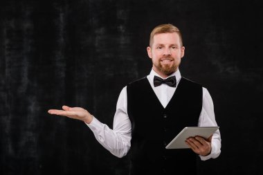 Happy young bearded waiter with digital tablet showing you where to go while standing in front of camera clipart